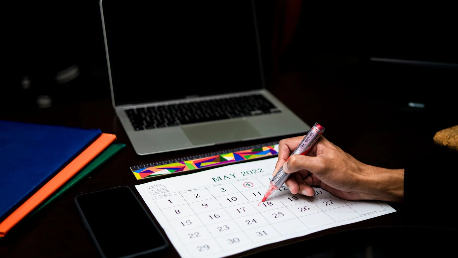 Hand circling a date on a paper desk calendar next to a closed laptop and colored folders