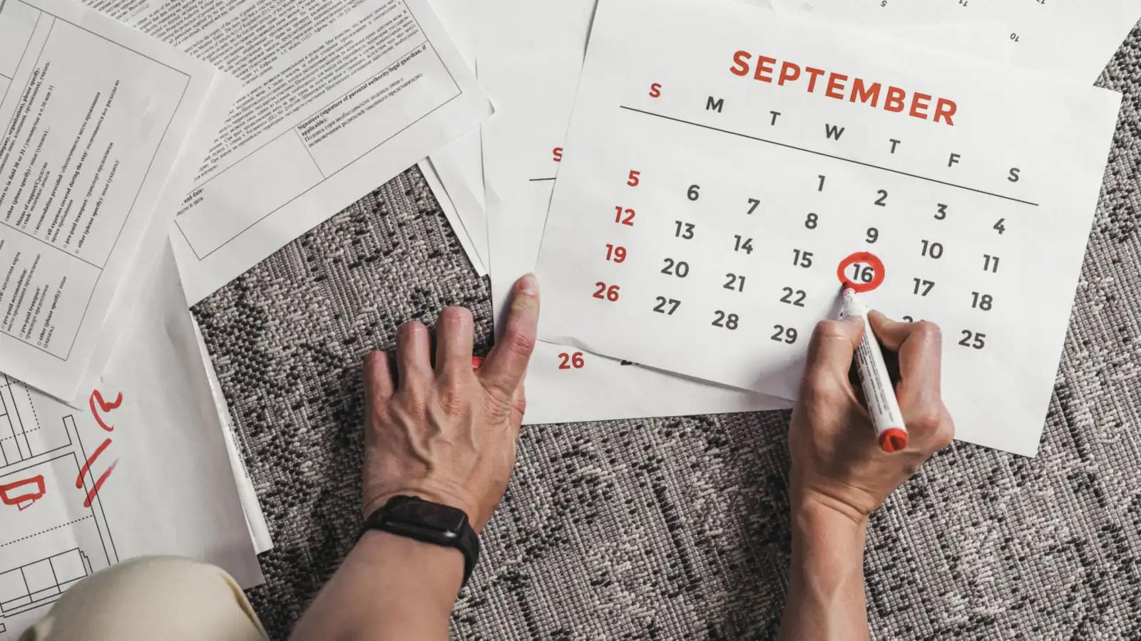 Hands marking a date with a red marker on a paper calendar, with printed forms spread across the floor