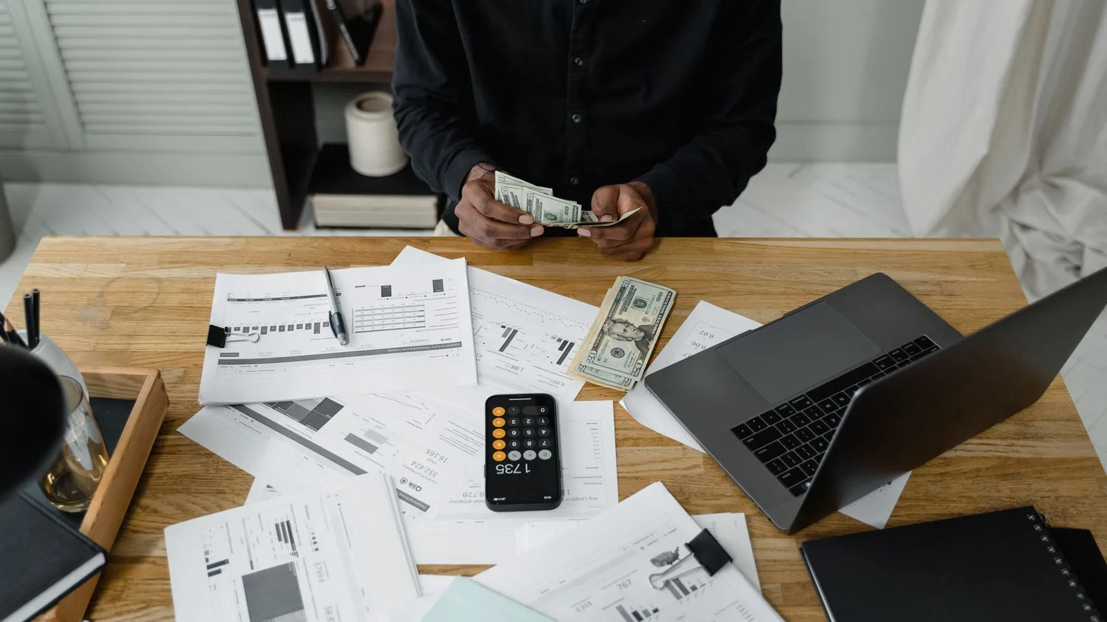 Hands counting dollar bills beside financial records and a laptop
