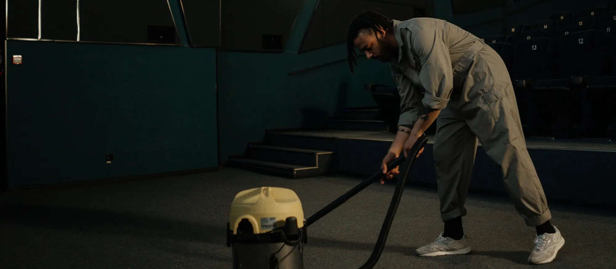 Cleaner in gray coveralls vacuuming the carpet of an empty movie theater with a wet-dry vacuum