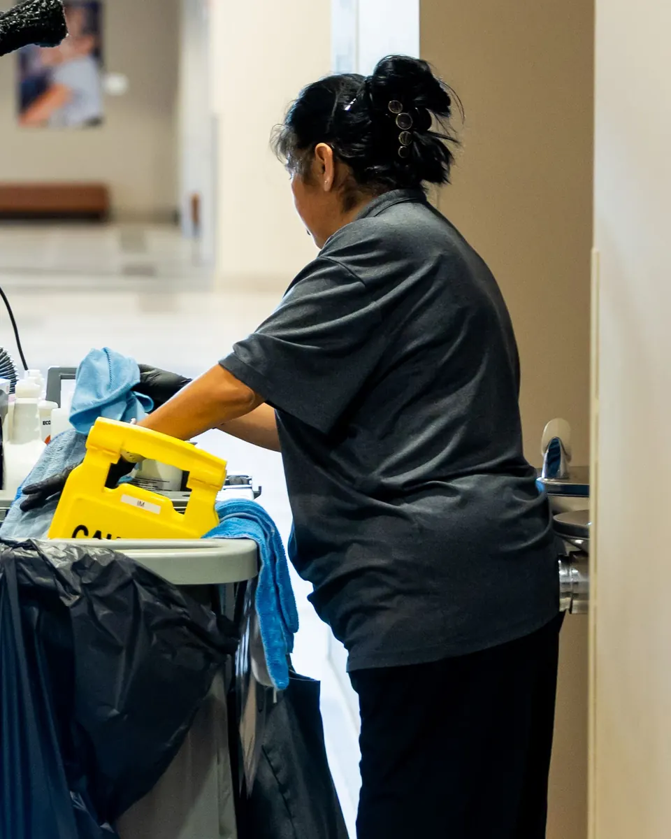 Janitorial worker in a gray uniform shirt reaching into her cleaning cart in a commercial building hallway