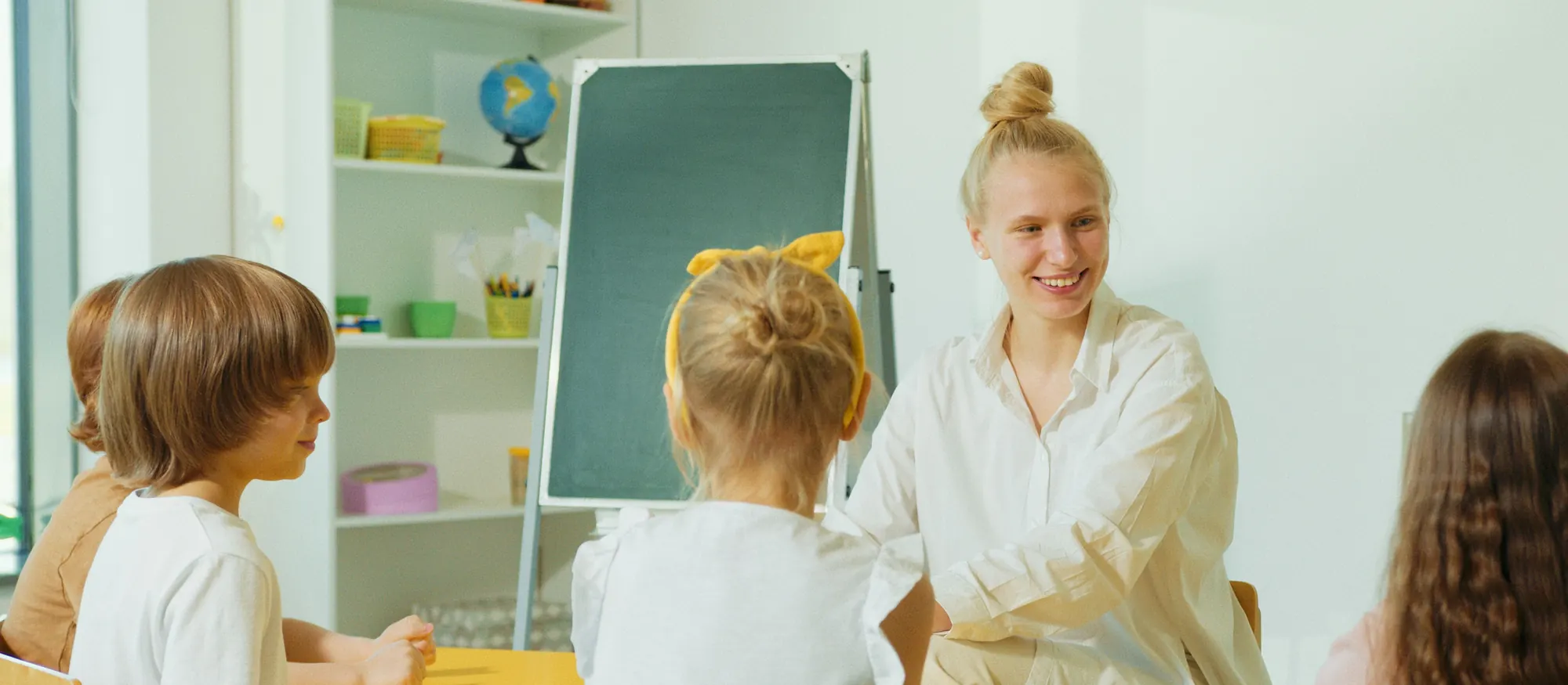 Smiling child care teacher seated at a yellow table with preschoolers seen mostly from behind