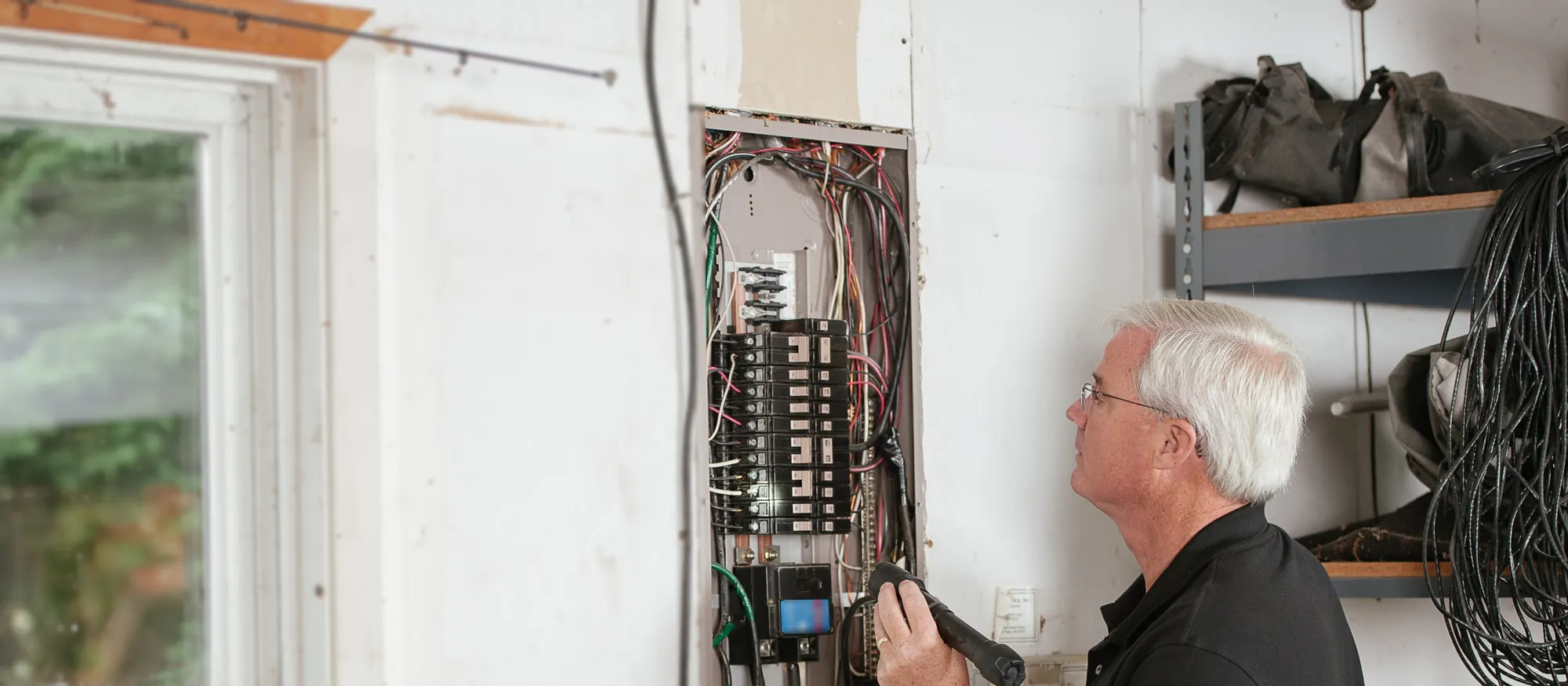 Electrician with a flashlight inspecting the wiring of a residential breaker panel in a garage