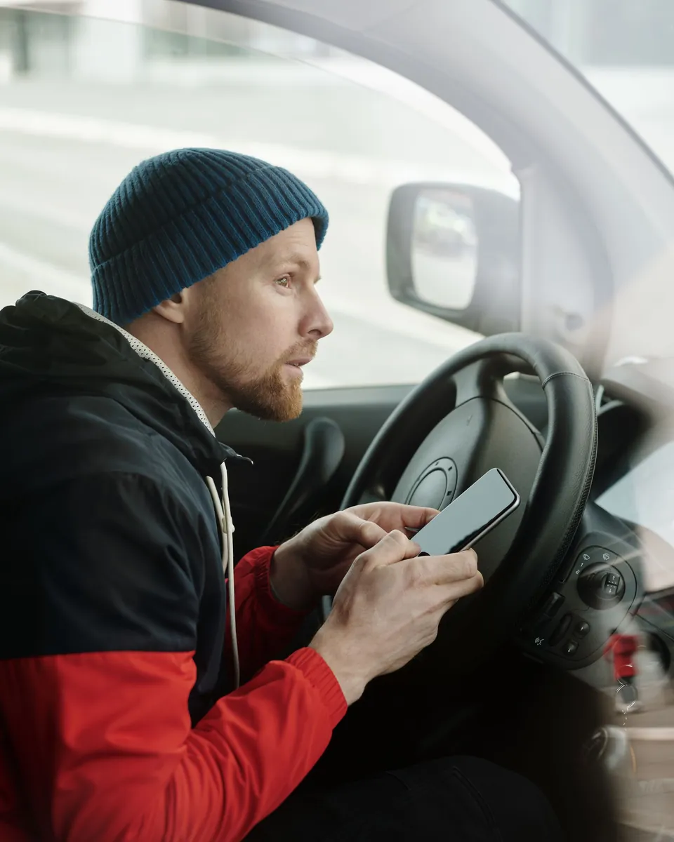 Driver in a knit cap checking his phone behind the wheel of his car between trips