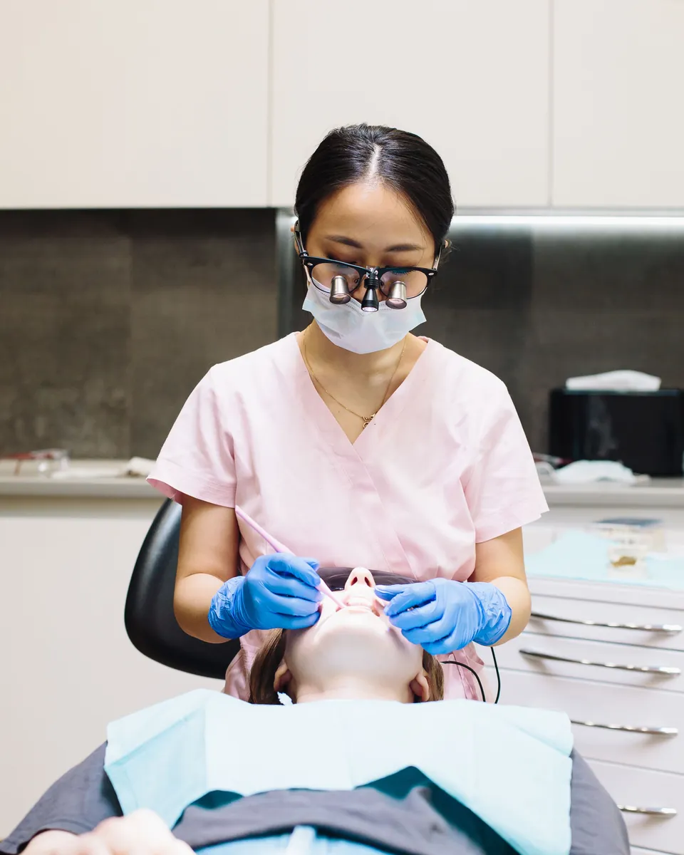 Dentist in pink scrubs and magnifying loupes examining a patient's teeth in her practice
