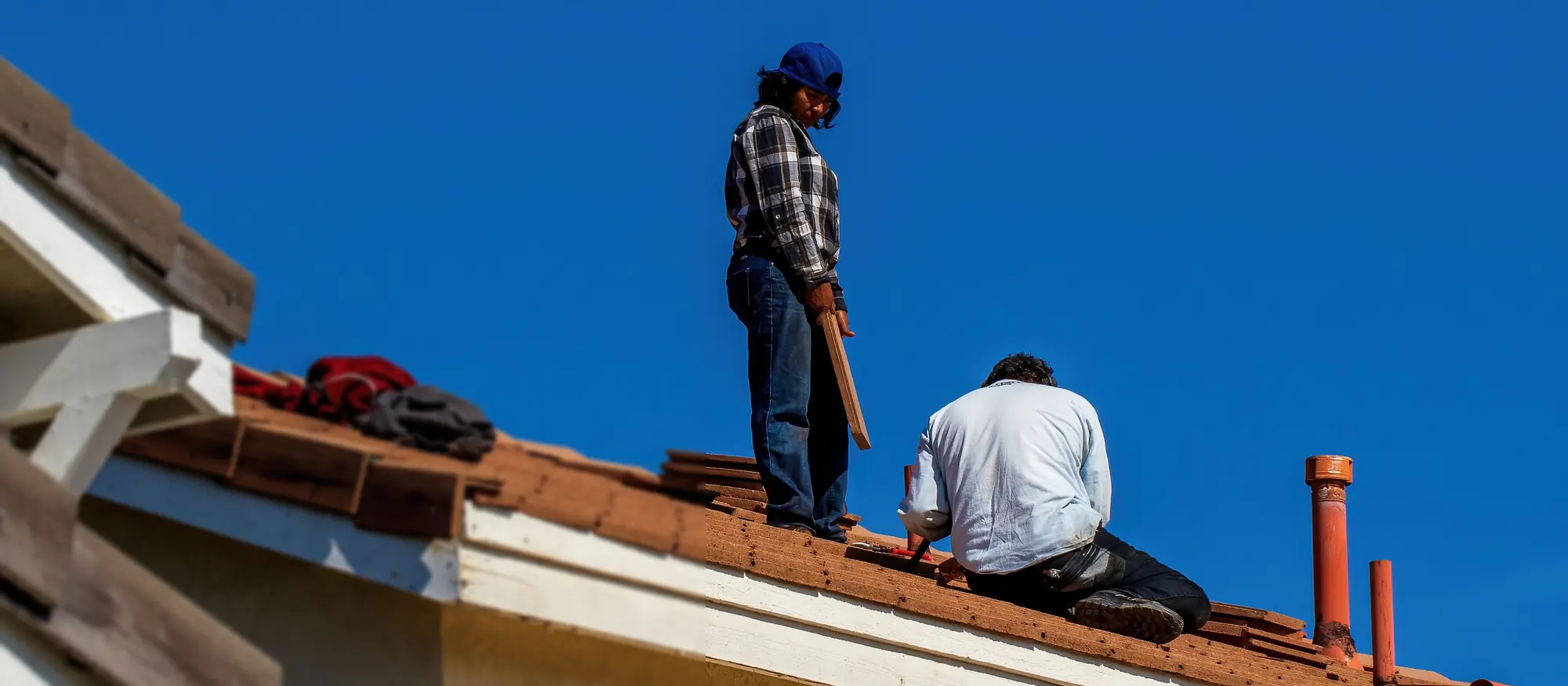 Two roofers replacing clay tiles on a stucco house under a clear blue sky