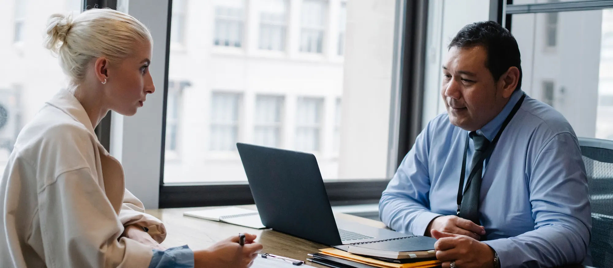 Recruiter in a blue shirt and lanyard interviewing a job applicant who fills out a form at a desk by the window