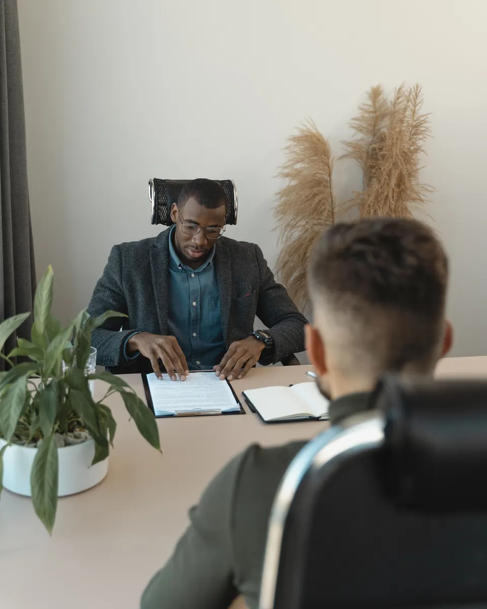 Recruiter in glasses reading a candidate's application on a clipboard while the applicant sits across the desk
