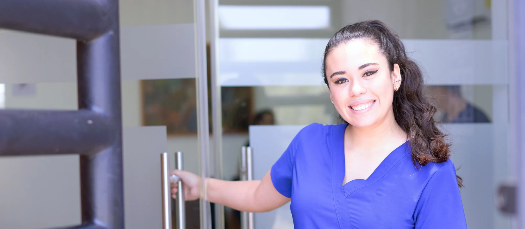 Smiling nurse in royal blue scrubs holding open the glass door of a clinic