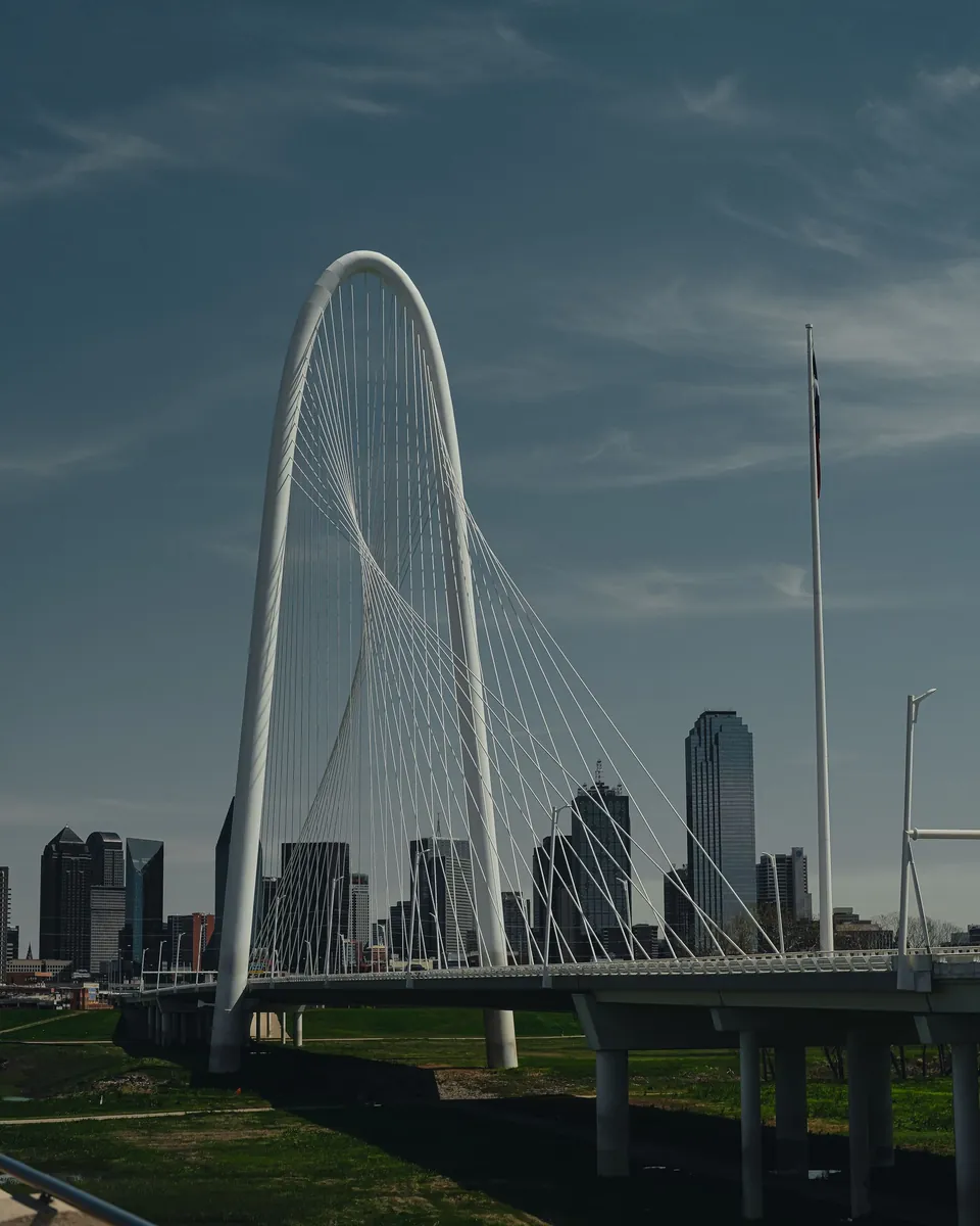 White arch of the Margaret Hunt Hill Bridge with the downtown Dallas, TX skyline behind it